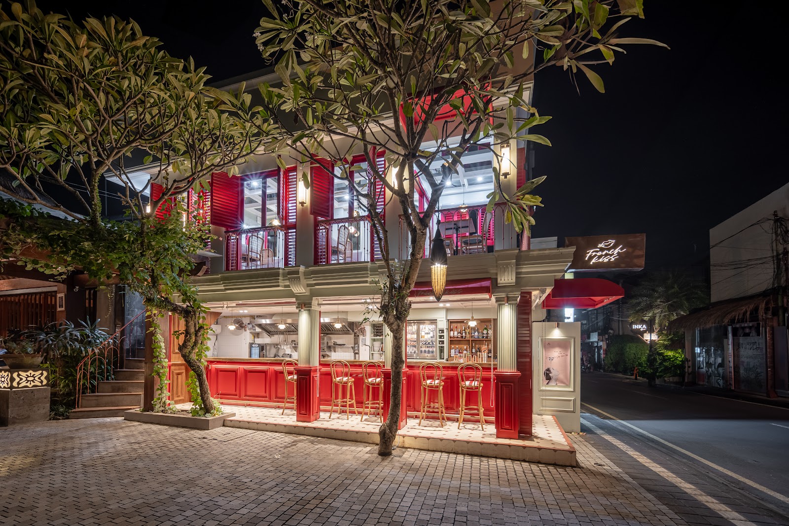 Night exterior view of a stylish restaurant or bar establishment with striking red shutters and doors, cream-colored architectural details, elegant lighting, and tropical flowering trees creating an upscale ambiance