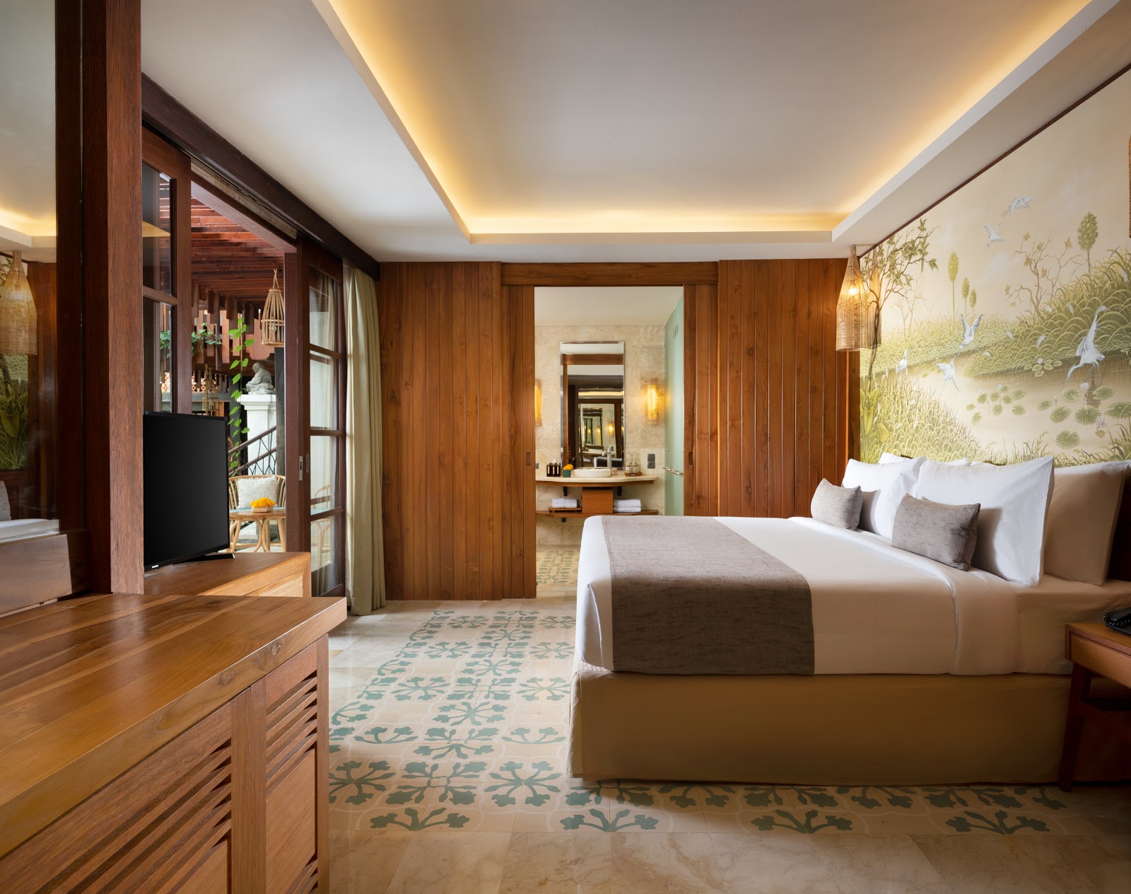 Luxurious bedroom suite featuring rich wood paneling, a king-size bed with premium bedding, decorative Asian-inspired artwork, ambient ceiling lighting, and an en-suite bathroom visible through open doorway. The room showcases elegant design with warm wood tones, patterned carpet, and modern amenities including a TV and climate control.