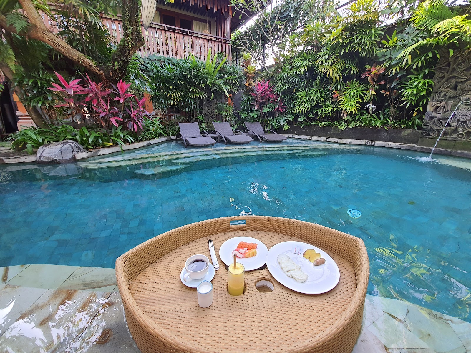 Luxurious swimming pool area with breakfast setup on woven round table in foreground. Crystal clear turquoise water surrounded by lush tropical vegetation, pink flowering plants, lounge chairs, and traditional Balinese architecture with wooden buildings and carved stone details in background.
