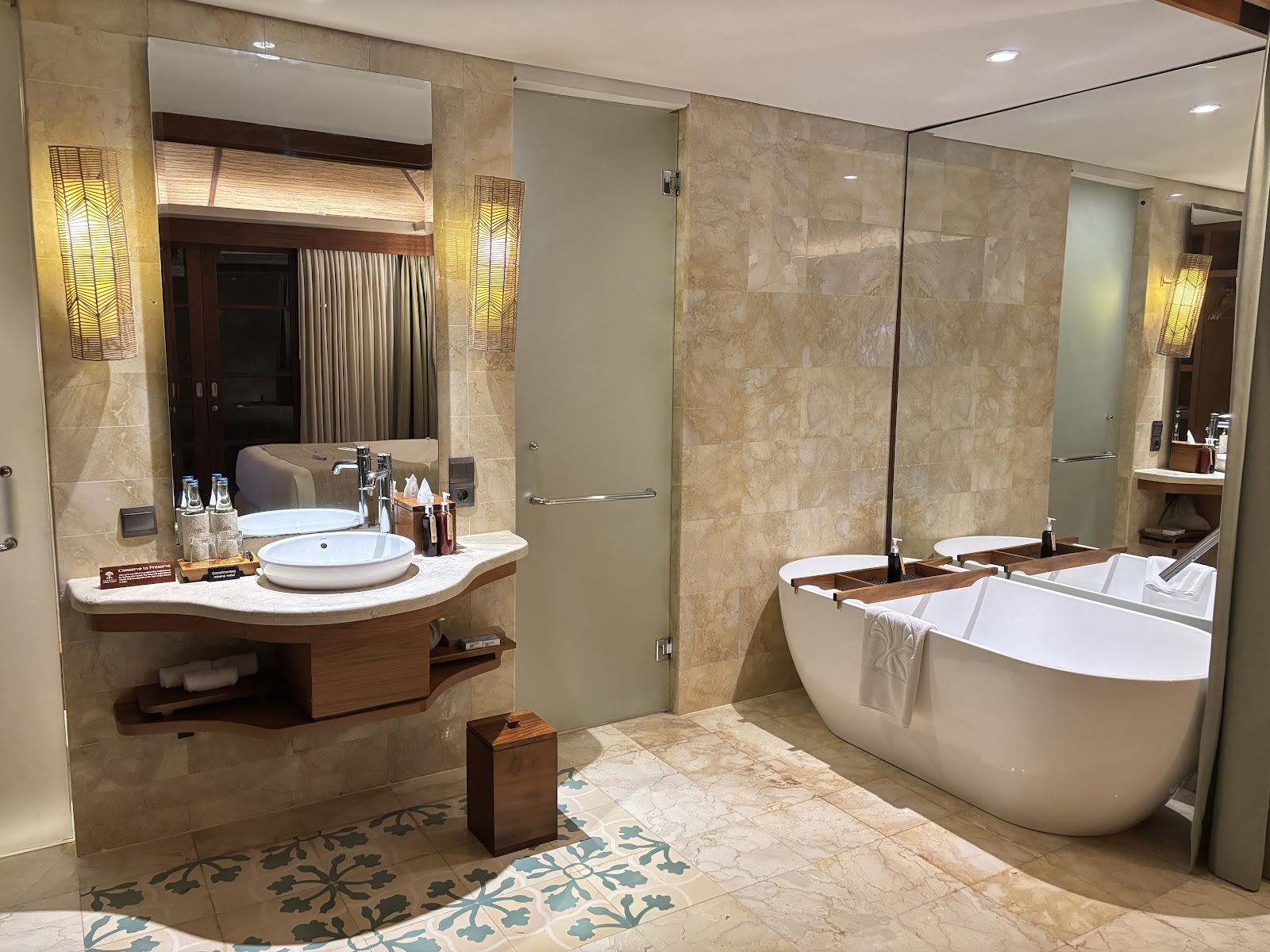 Luxurious bathroom featuring a large white freestanding bathtub, elegant curved sink vanity with round vessel basin, marble tile walls, contemporary lighting fixtures, and decorative floor patterns with a tropical color scheme.