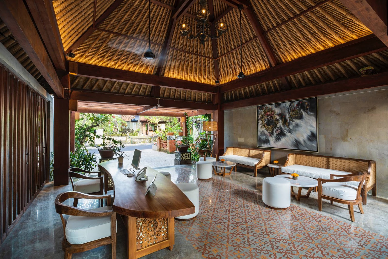 Elegant lobby or reception area with traditional Balinese architecture, featuring a stunning coffered ceiling with warm golden lighting, carved wooden furnishings, patterned floor tiles, and modern amenities. The space opens to a courtyard garden visible through large windows, combining contemporary comfort with cultural design elements.