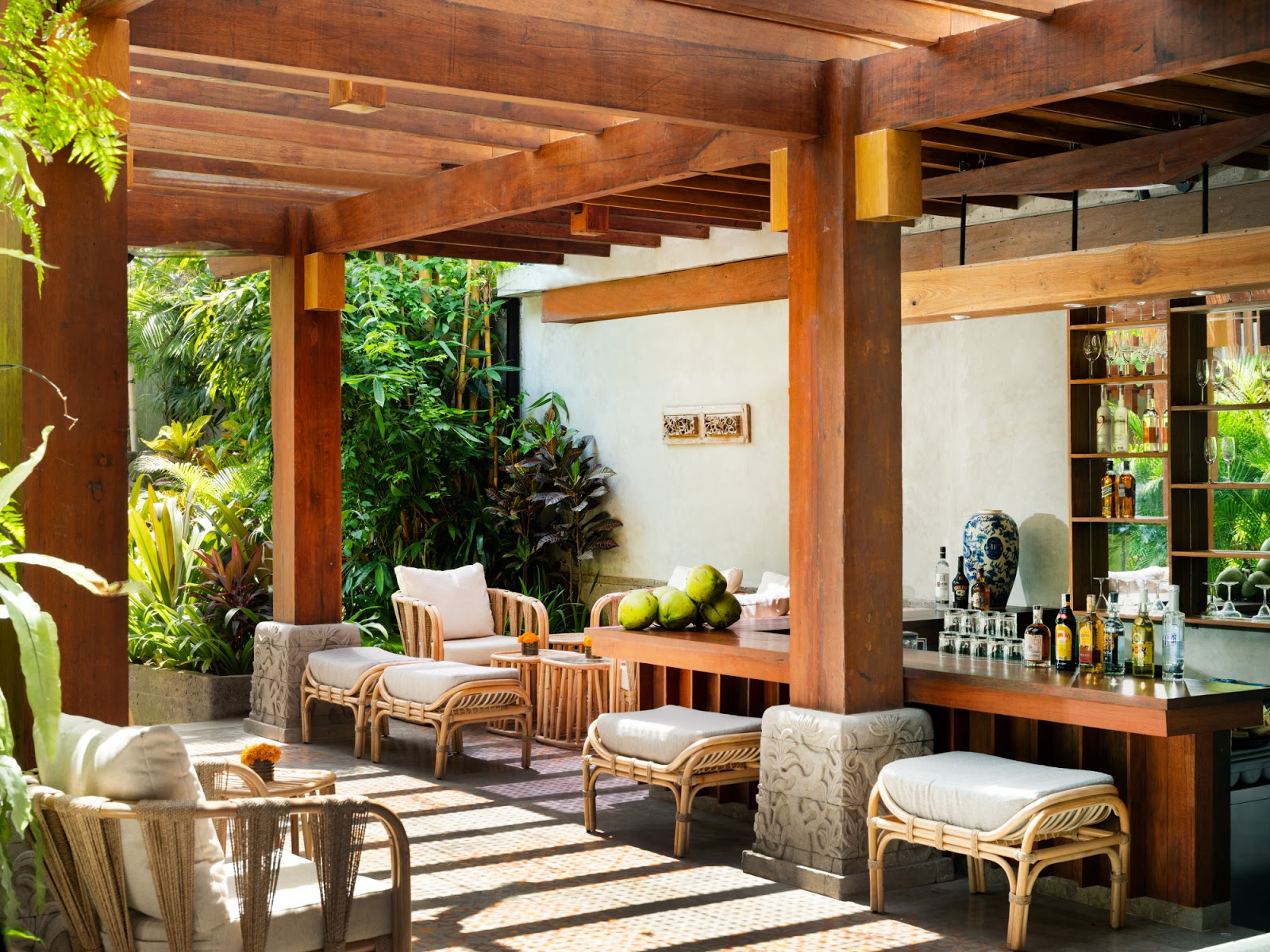 Luxurious covered outdoor lounge area with wooden pergola, rattan furniture with white cushions, built-in bar counter, and lush tropical greenery. Professional resort design with excellent natural lighting and sophisticated interior-exterior integration.
