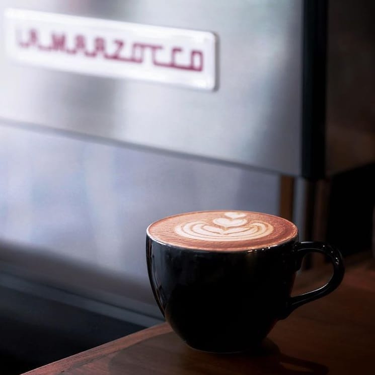 A beautifully crafted latte with latte art in a black cup, placed on a wooden surface with a blurred 'Unmarked' sign visible in the background, suggesting an intimate café setting